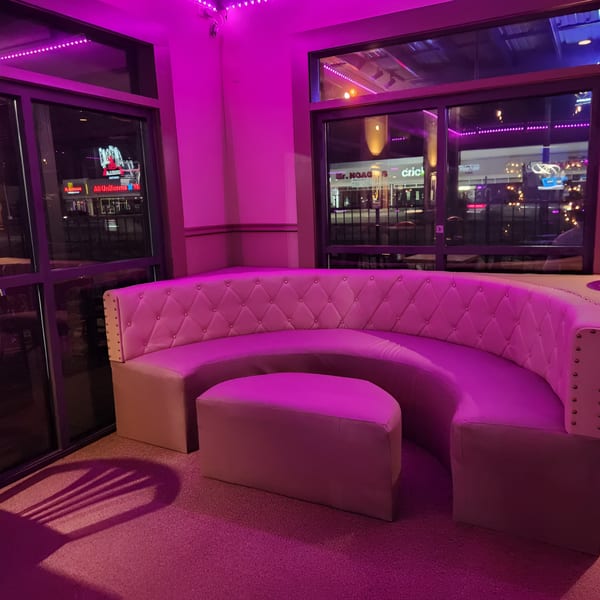 Curved pink booth seating with a matching ottoman under purple lighting at Shades Bar & Grill.