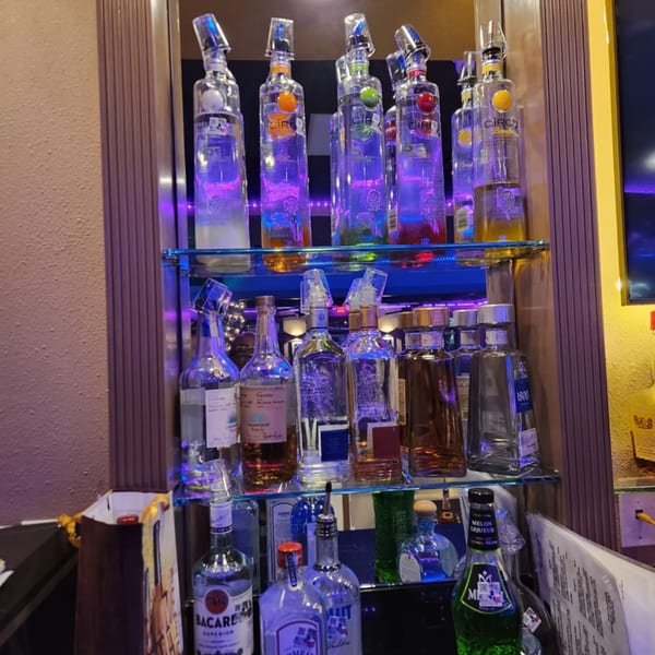 A bar shelf at Shades Bar & Grill displaying various liquor bottles, illuminated by purple and blue lights.