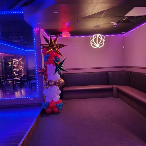 A lounge area with purple and blue lighting, a balloon column, and brown L-shaped booth seating.