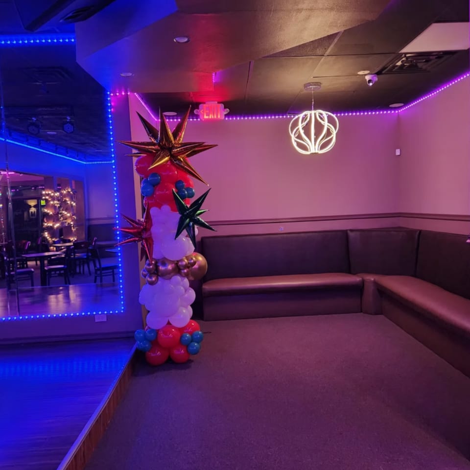 A lounge area with purple and blue lighting, a balloon column, and brown L-shaped booth seating.